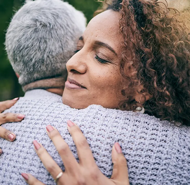 Two people hugging outdoors, one facing away with gray hair, the other with curly hair and closed eyes, smiling contentedly.