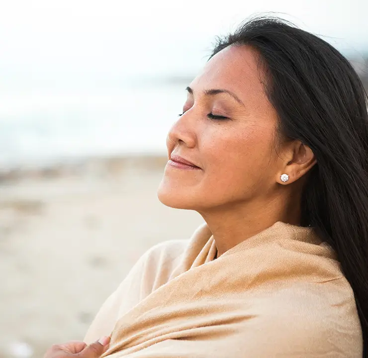 A woman with closed eyes smiles gently, standing on a beach with the ocean in the background.