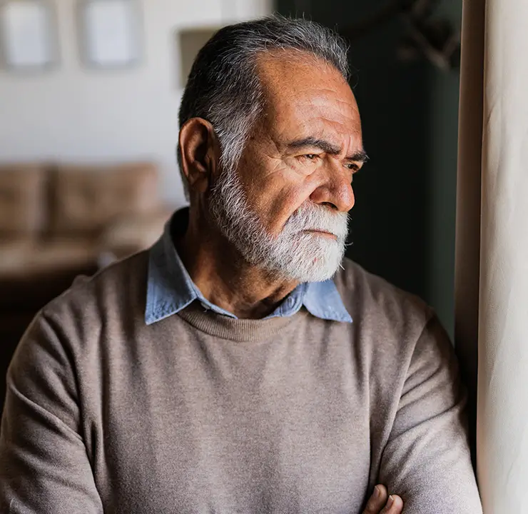 Elderly man with a beard gazes thoughtfully out a window in a cozy living room.