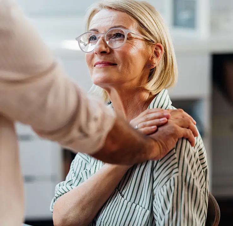 An older woman wearing glasses looks up, smiling, while a person stands nearby with a hand on her shoulder.