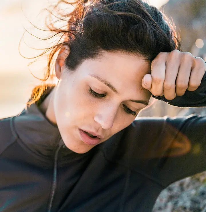 A woman in a black jacket appears tired, resting her forehead on her hand outdoors, with sunlight in the background.