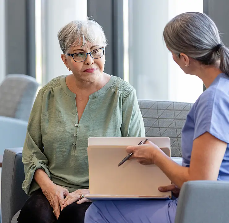 A nurse in scrubs talks to an older woman with glasses sitting on a chair in a waiting area near large windows.