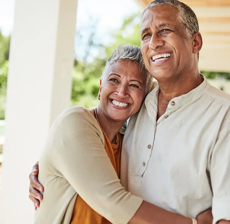 Smiling couple embracing outdoors on a sunny day, standing on a porch with greenery in the background.