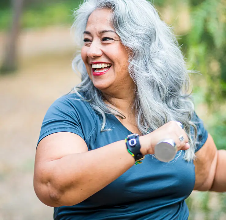 A smiling woman with long gray hair stands outdoors, wearing a blue shirt, against a blurred green background.