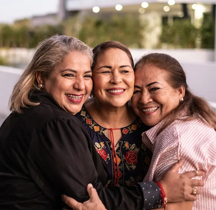 Three women smiling and embracing outdoors, with a pool and trees in the background.
