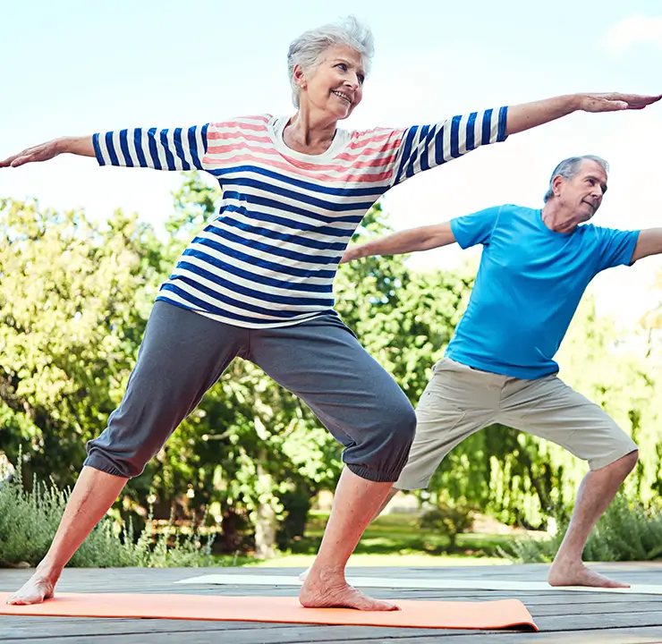 Two older adults practice yoga outdoors, standing in a warrior pose. They are surrounded by greenery and a clear blue sky.