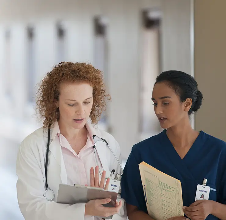 Two medical professionals, one in a white coat and the other in blue scrubs, discuss notes in a hallway.