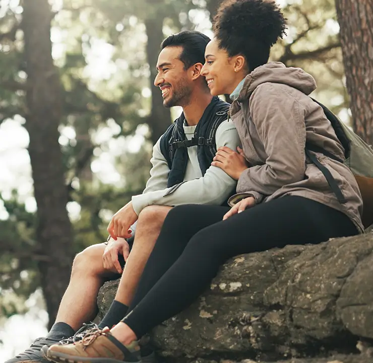 A couple sits on a rock, smiling and enjoying the forest view, surrounded by tall trees and sunlight filtering through the leaves.