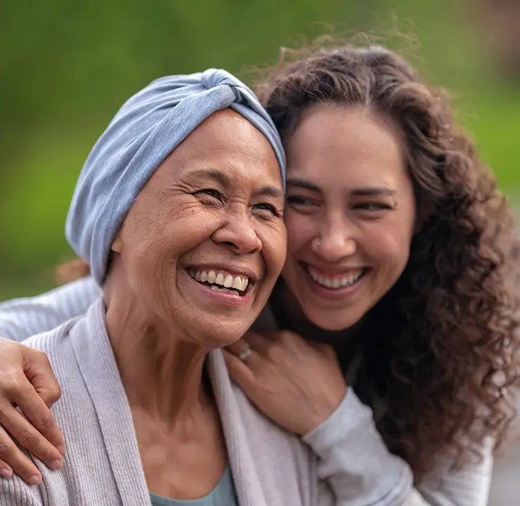 Elderly woman with a headscarf smiles as a younger woman with curly hair hugs her from behind in a park.