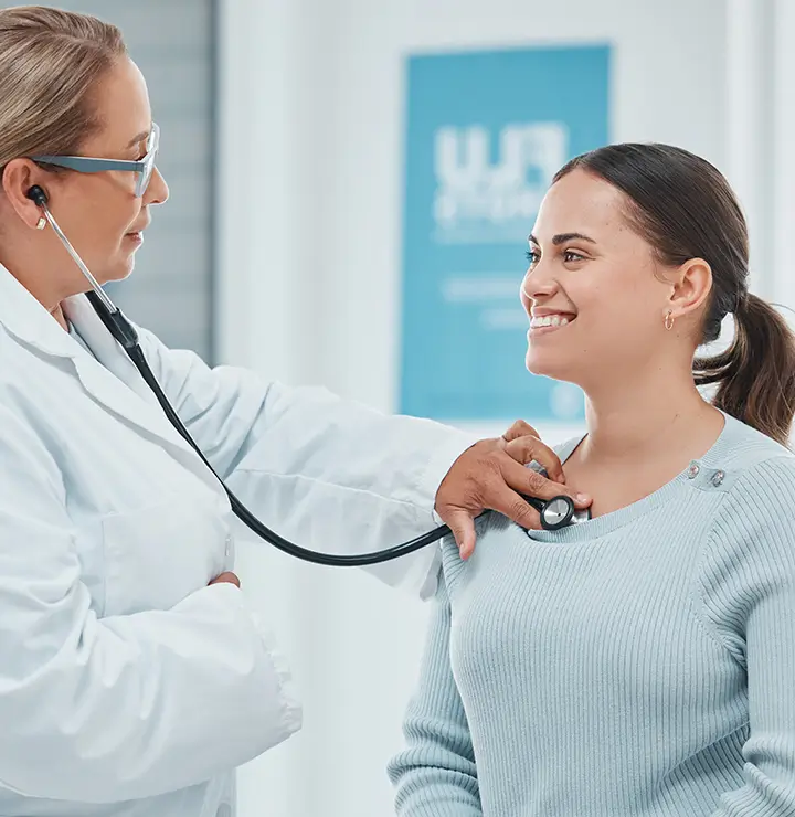 A doctor uses a stethoscope to examine a smiling patient in a medical office.