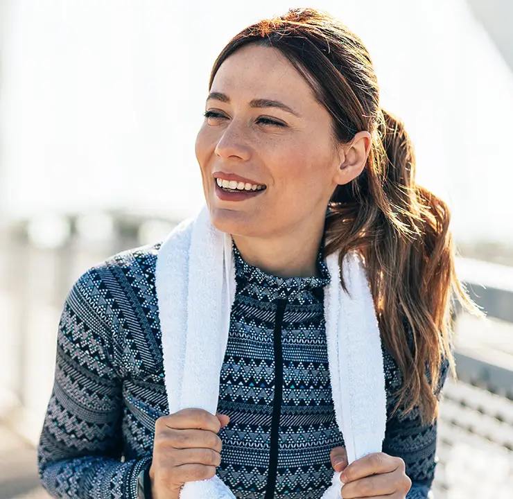 Woman with a towel around her neck, smiling, standing outside on a sunny day with a bridge in the background.
