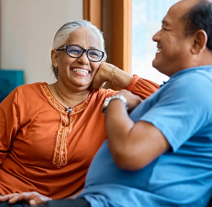 An elderly couple smiling and conversing in a cozy room with shelves filled with books in the background.