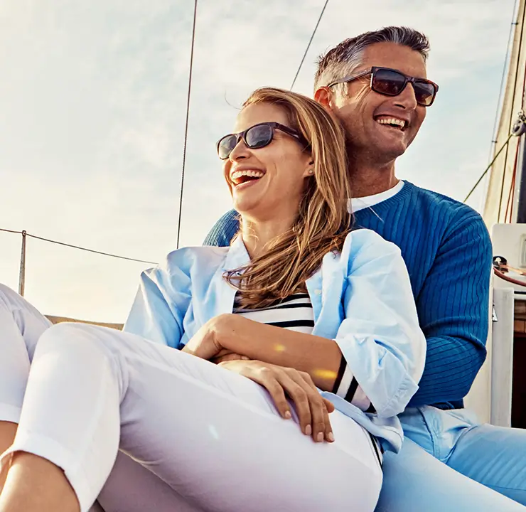 A couple wearing sunglasses smiles while sitting on a boat under a sunny sky.