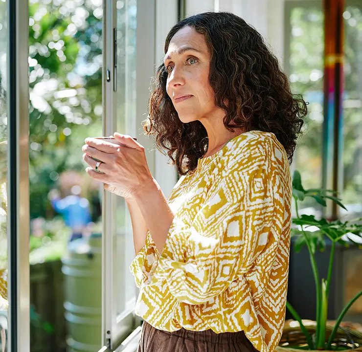 Woman with curly hair, wearing a yellow patterned shirt, holding a cup and gazing out a window, with greenery visible outside.