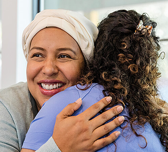 Two people hugging on a couch, one wearing a headscarf and smiling warmly, the other with curly hair facing away.