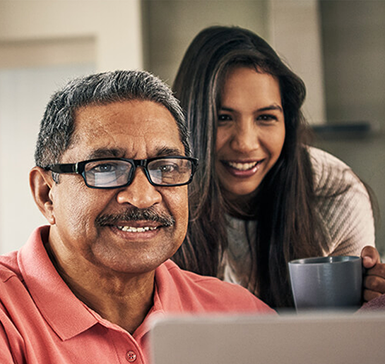 Three people smiling and looking at a laptop screen. The older man and woman are seated, and the younger woman stands behind them holding a mug.