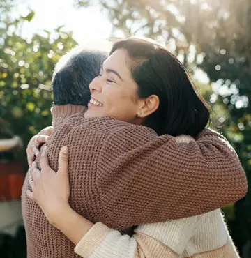 A woman and a man hug outside, smiling, with sunlight filtering through the trees in the background