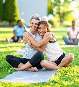 A white middle aged woman and a young Asian woman are smiling as they hug in an outdoor yoga class.